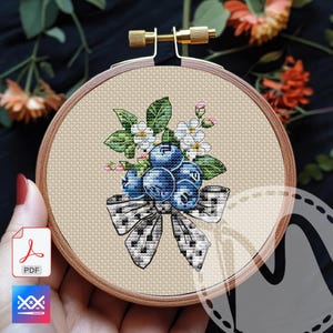 Könnte beinhalten: Ein Kreuzstich-Stickrahmen mit einem Design aus Blaubeeren, weißen Blüten und grünen Blättern. Unten befindet sich eine schwarz-weiße Schleife. Die Stickerei befindet sich in einem Holzrahmen mit einer Messing-Einstellschraube.