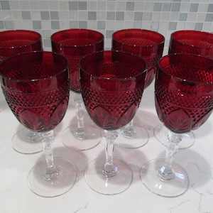 Puede incluir: Conjunto de ocho copas de vino tinto rubí. Cada copa presenta un patrón de corte de diamante en el cuenco, con un tallo y una base transparentes. Las copas están dispuestas sobre una superficie blanca, mostrando su color y diseño.