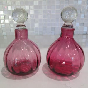 Puede incluir: Dos frascos de perfume antiguos de cristal rosa con tapones redondos y transparentes. Los frascos tienen un diseño acanalado y una forma bulbosa. Los tapones son esféricos. Los frascos se exhiben sobre una superficie blanca.