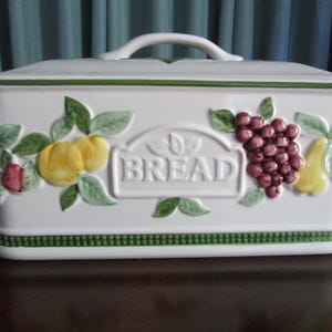 May include: White ceramic bread box with a handle on the lid. The box is decorated with embossed fruit and the word "BREAD". Green trim and accents add to the design. The bread box is rectangular.