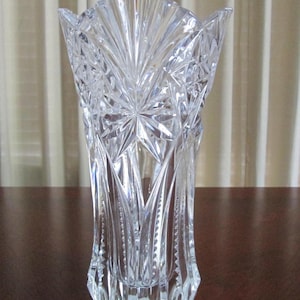 Könnte beinhalten: Eine transparente, geschliffene Glasvase mit ausgestellter Oberseite und geometrischem Muster. Die Vase hat eine hohe, schlanke Form und steht auf einer dunklen Holzoberfläche. Das geschliffene Glasdesign erzeugt einen funkelnden Effekt und reflektiert das Licht.