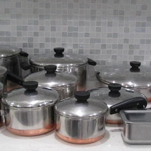 May include: A collection of stainless steel cookware featuring copper-toned bases and black handles. The set includes various pots, pans, and lids of different sizes, along with a rectangular metal container, all arranged on a white surface.