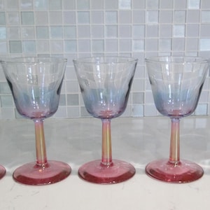 May include: Five stemmed wine glasses with a pink base and a clear bowl that fades to blue. The stems are a light gold color. The glasses are arranged in a row.