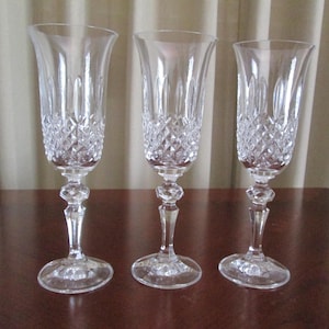 May include: Three clear crystal champagne flutes featuring a diamond-cut pattern on the lower bowl, vertical fluting near the rim, and a faceted, multi-sided stem leading to a circular base. The glasses are transparent with reflective surfaces.