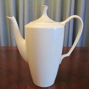 May include: A tall, white porcelain coffee pot with a slender spout and a curved handle. The lid is topped with a small, rounded finial. Gold trim accents the spout, lid, and handle, adding a touch of elegance to the design. The pot sits on a dark wooden surface.