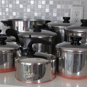 May include: A collection of stainless steel cookware featuring multiple pots and pans with copper-colored bases and black handles. Each piece includes a matching lid with a black knob. The set is arranged in a group on a flat surface.