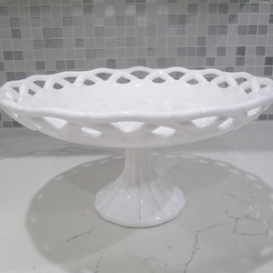 Puede incluir: Un soporte para pasteles blanco con diseño de celosía en el borde. El soporte tiene una base redonda y un tallo acanalado. El soporte para pasteles está hecho de cerámica o porcelana y es adecuado para exhibir postres.