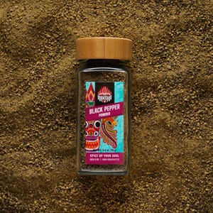 May include: A clear glass jar of black pepper powder with a wooden lid. The label features the text "RAKSHA BLACK PEPPER POWDER" and a colorful illustration. The jar contains 70 g of black pepper.