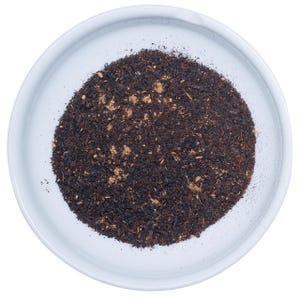 May include: A white ceramic bowl filled with a mound of loose-leaf black tea. The tea consists of dark, finely processed leaves mixed with small, light brown particles and fibrous bits, creating a textured, granular appearance within the bowl.