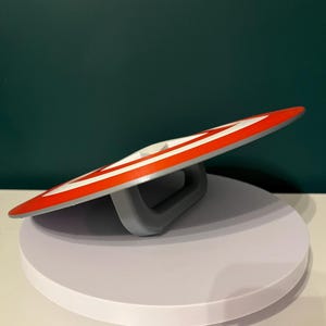 May include: A circular shield in red, white, and grey, featuring a striped pattern and a central white star. The shield has a grey handle on the back. The design is reminiscent of a superhero's emblem.