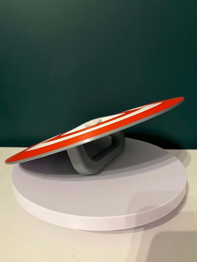 May include: A circular shield in red, white, and grey, featuring a striped pattern and a central white star. The shield has a grey handle on the back. The design is reminiscent of a superhero's emblem.