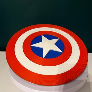 May include: A circular shield in red, white, and blue, featuring a white star at the centre. The design includes a red outer ring, a white middle ring, and a blue central area. This shield is a replica of Captain America's iconic shield.