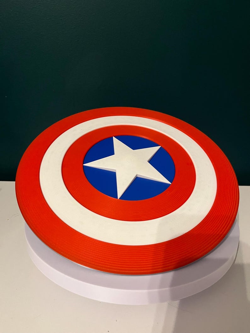 May include: A circular shield in red, white, and blue, featuring a white star at the centre. The design includes a red outer ring, a white middle ring, and a blue central area. This shield is a replica of Captain America's iconic shield.