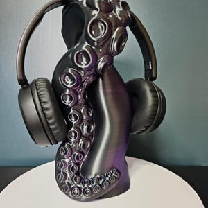 Può includere: Un supporto per cuffie a forma di tentacolo di polpo nero e viola. Il supporto tiene un paio di cuffie nere over-ear. Il tentacolo ha una superficie strutturata con dettagli di ventose. Il supporto è su una superficie bianca.