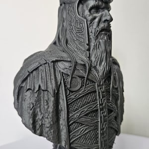 Può includere: Busto grigio scuro di un re con corona e armatura dettagliate. La scultura presenta una lunga barba e intagli intricati, con una base ornata di teschi. Il design generale suggerisce un tema fantasy o storico.