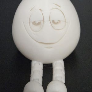 Könnte beinhalten: Ein weißes, eiförmiges Spielzeug mit einem lächelnden Gesicht und zwei Beinen. Das Spielzeug hat ein einfaches Design und ist aus glattem, weißem Material gefertigt.