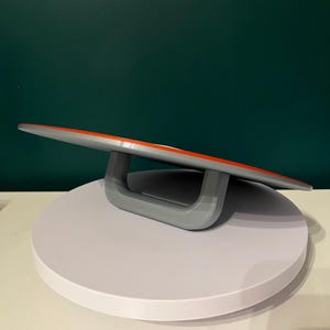 May include: A grey and orange serving board with a curved design, supported by a grey handle. The board is set on a white, circular platform, with a dark green background.