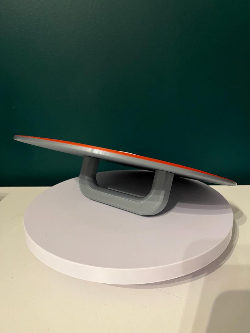 May include: A grey and orange serving board with a curved design, supported by a grey handle. The board is set on a white, circular platform, with a dark green background.