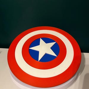 May include: A circular shield in red, white, and blue, featuring a white star at the centre. The design includes a red outer ring, a white inner ring, and a blue central area. This shield is a replica of Captain America's iconic shield.
