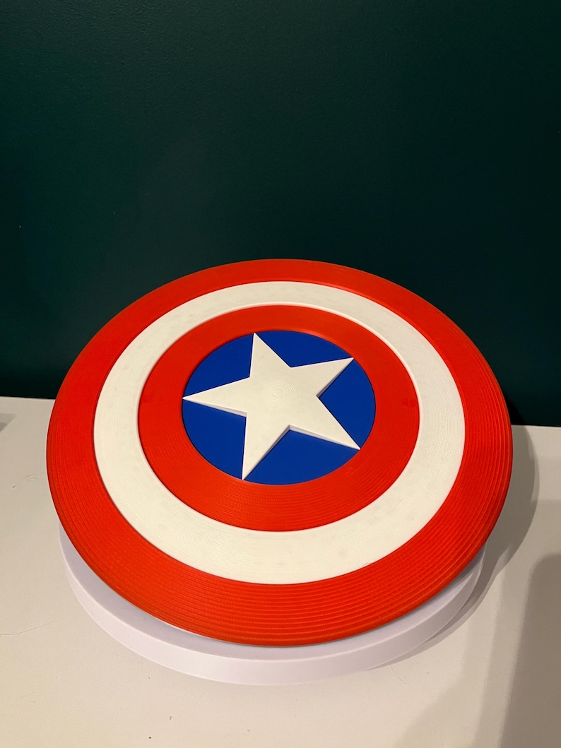 May include: A circular shield in red, white, and blue, featuring a white star at the centre. The design includes a red outer ring, a white inner ring, and a blue central area. This shield is a replica of Captain America's iconic shield.