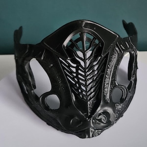 May include: A black, intricately designed mask with a textured surface. The mask features a central, stylized design with a series of vertical lines and a mesh-like pattern. The mask has openings for the eyes and mouth, and curved extensions on the sides.