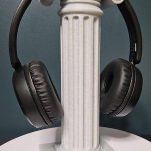 Puede incluir: Auriculares negros sobre un soporte decorativo en forma de columna clásica, de color gris claro. El soporte tiene un diseño acanalado y una base cuadrada. Los auriculares tienen almohadillas acolchadas.