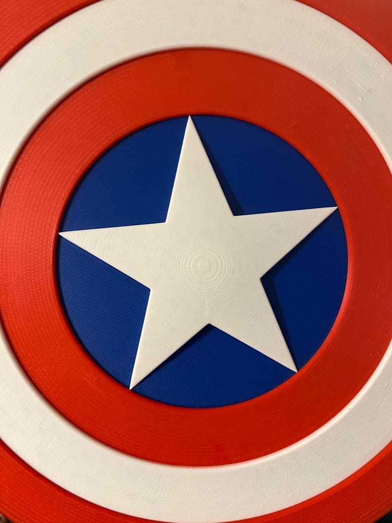 May include: A shield in red, white, and blue, featuring a white star at its centre. The design is circular, with a white outer ring, a red middle ring, and a blue centre containing a white star.