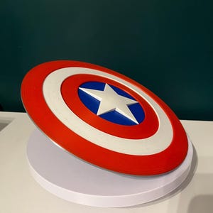 May include: A circular shield in red, white, and blue, featuring a white star in the centre. The shield has a red outer ring, a white middle ring, and a blue central area. This is a prop or collectible item.