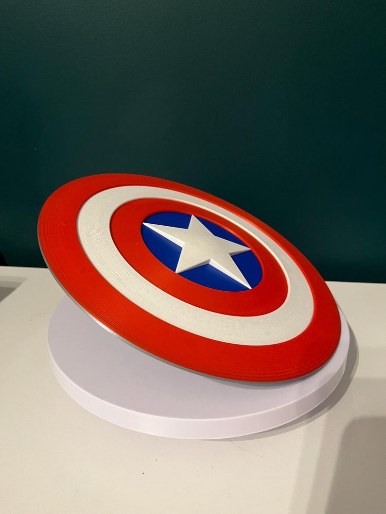 May include: A circular shield in red, white, and blue, featuring a white star in the centre. The shield has a red outer ring, a white middle ring, and a blue central area. This is a prop or collectible item.