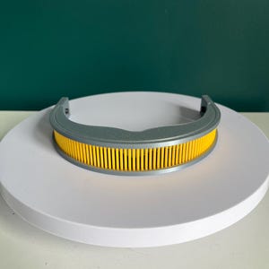 Puede incluir: Un filtro de aire gris y amarillo. El filtro tiene una forma curva con un marco gris y un interior amarillo y acanalado. El objeto está sobre una superficie blanca y un fondo verde oscuro.