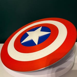 May include: A circular shield in red, white, and blue, featuring a white star at the centre. The design includes concentric rings of red, white, and red, with a blue central circle. It is a replica of Captain America's shield.
