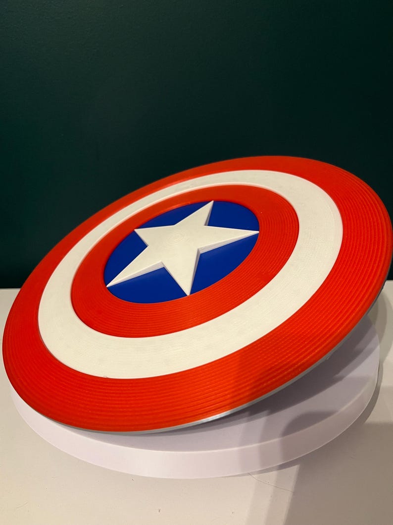 May include: A circular shield in red, white, and blue, featuring a white star at the centre. The design includes concentric rings of red, white, and red, with a blue central circle. It is a replica of Captain America's shield.
