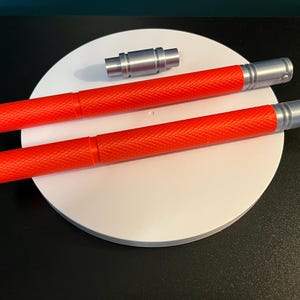 May include: Two red, textured, cylindrical objects with silver ends and a small silver cylindrical piece are displayed on a white circular surface. The objects have a diamond pattern.