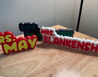 May include: Three-dimensional name signs in red, yellow, white, and green. The signs read "MRS. MAY", "MRS. BOOK", and "MRS. BLANKENSHIP". The signs are on a wooden surface.