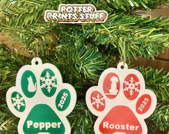 May include: Christmas tree ornaments shaped like paw prints. One is green with a cat silhouette, snowflakes, and the name "Pepper." The other is red with a dog silhouette, snowflakes, and the name "Rooster." Both have the year "2025."