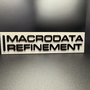 May include: White rectangular sign with black text that reads "MACRODATA REFINEMENT".