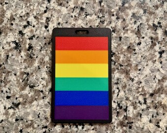 May include: A rectangular black luggage tag with a rainbow design. The rainbow stripes include red, orange, yellow, green, blue, and purple. The tag has a small hole at the top for attachment.