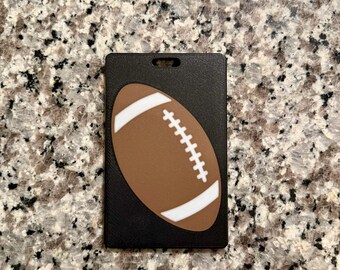 May include: A black luggage tag with a brown and white football design. The football has white stripes and stitching. The tag has a slot for attaching to luggage or a bag.