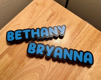 May include: Two custom name signs, "Bethany" and "Bryanna", in a bubble font. The names are in light blue with a black outline, set against a wooden background. These personalized signs are ideal for room decor.
