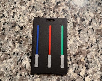 May include: A black luggage tag featuring three lightsabers in blue, red, and green. The lightsabers have white handles. The tag has a hole at the top for attachment.