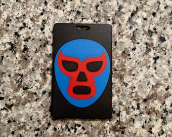 May include: Black rectangular luggage tag featuring a blue mask with red accents. The mask design is centered on the tag, which has a small hole at the top for attachment. The tag is likely made of a durable material.