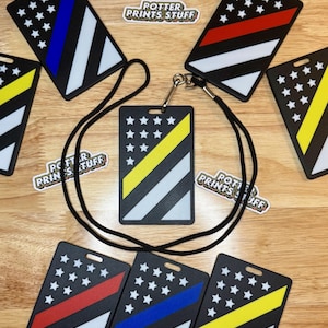 May include: A set of six black plastic ID badge holders with a thin blue, red, or yellow stripe and a white star pattern. The badge holders are on a wooden surface and have a black lanyard.