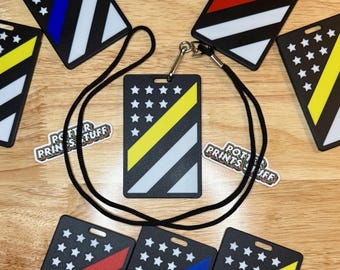 May include: Black ID badge holders with various flag-inspired designs. Each features a diagonal stripe in colors like blue, red, and yellow, along with white stripes and a field of white stars on a black background. A black lanyard is attached to one.