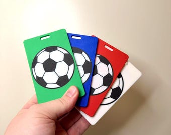 May include: A hand holding four rectangular soccer-themed tags in green, blue, red, and white. Each tag features a black and white soccer ball design. The tags have a small hole at the top for attachment.