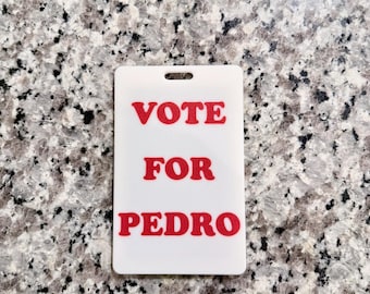 May include: A white rectangular badge with a hole at the top. The badge has the words "VOTE FOR PEDRO" printed in red, bold letters. The background is a speckled gray and white surface.