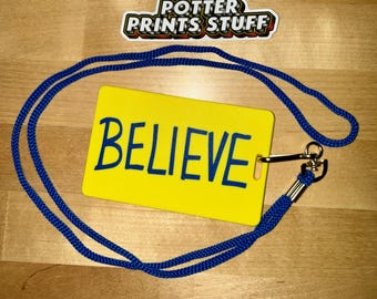 May include: A yellow rectangular tag with the word "BELIEVE" in dark blue lettering, attached to a blue lanyard. The tag is centered, with the lanyard looped around it. The "Potter Prints Stuff" logo is in the upper right corner.