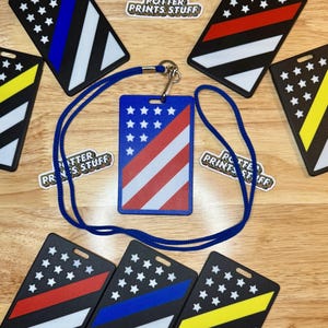 May include: A set of six American flag themed ID badge holders with a blue lanyard. The badge holders feature a black background with a white stripe and a red, blue, or yellow stripe. The lanyard is made of a blue cord.