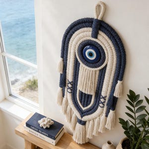 May include: A wall hanging featuring a central blue and white glass evil eye charm surrounded by intricate navy blue and cream macrame cord patterns, including decorative tassels and cross-stitch details, suspended by a sturdy top loop.
