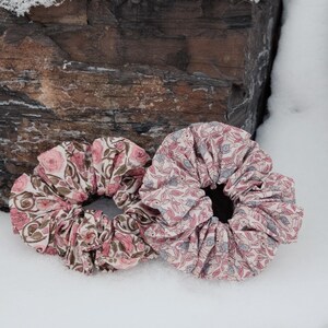 Puede incluir: Dos scrunchies de tela con estampados florales. Un scrunchie es rosa y blanco con un estampado floral, el otro es rosa y verde con un estampado floral.