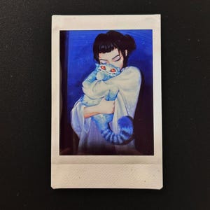 Op de afbeelding: Een Polaroid-achtige afdruk met een persoon die een blauwe kat omhelst. De kat heeft rode ogen en een gestreepte staart. De achtergrond is diepblauw en de persoon draagt een wit kledingstuk. De afdruk staat tegen een zwarte achtergrond.